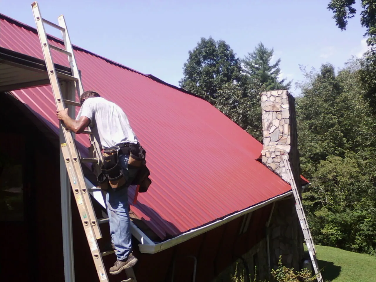 Roofer on a ladder beside a red metal roof and stone chimney — Soffit & Fascia Repair by Roofing Brothers in Haddam